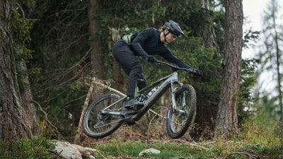 On voit un homme sur un vélo électrique Cube Stereo Hybrid gris qui saute par-dessus une pente dans la forêt.