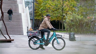 On voit un homme sur un vélo électrique de cargo, le Cube Longtail Hybird bleu qui roule dans le centre-ville avec des provisions.