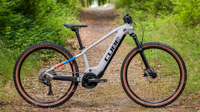 On voit un vélo électrique blanc pour enfants Cube, avec de petits détails bleus et rouges. Il se trouve sur un chemin forestier envahi par la végétation.