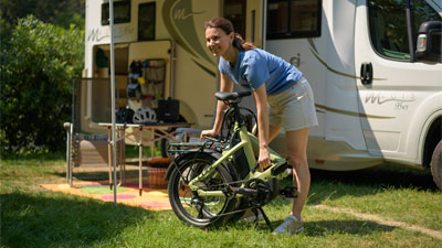 On voit une femme debout devant un camping-car. Devant elle se trouve un vélo pliant vert replié.