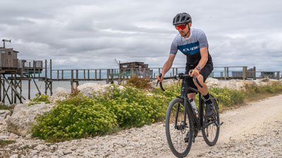 COn voit un homme en tenue de cycliste. Il roule sur son vélo de gravel sur une piste cyclable le long d'une plage.