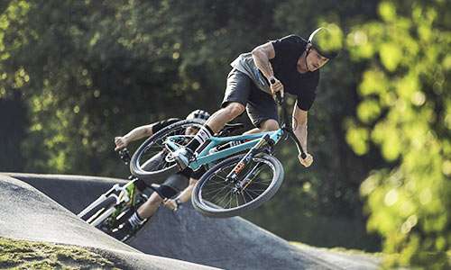 Un cycliste saute avec un vélo tout-terrain bleu turquoise dans un parc à vélos. À l'arrière-plan, on aperçoit un autre cycliste avec un vélo vert.