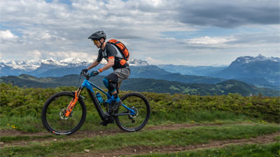 On voit un homme sur un VTT électrique Cube Stereo Hybrid bleu avec des détails orange. Il roule sur un sentier de randonnée avec des montagnes en arrière-plan.