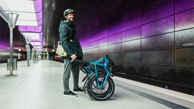 On voit un homme debout dans une station de métro. Devant lui se trouve un vélo pliant bleu replié.