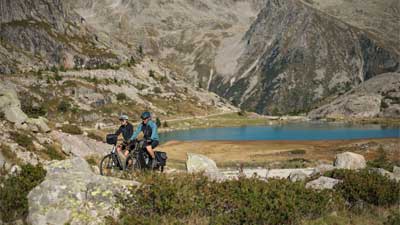 Un couple roule à vélo tout terrain dans un paysage rocailleux, avec des montagnes et un lac en arrière-plan.