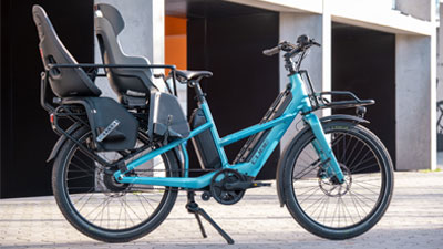 On peut voir un vélo de cargo, le Cube Longtail Hybrid bleu, équipé de deux sièges enfants. 