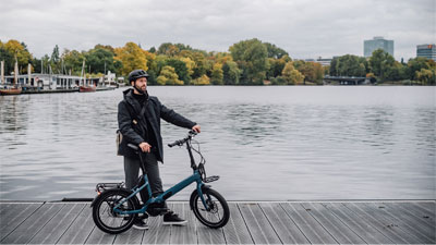 Un homme se tient à côté de son vélo pliant bleu sur une passerelle. À l'arrière-plan, on aperçoit un plan d'eau et une ville.