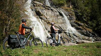 Un couple pousse ses vélos à travers un pré, avec une cascade en arrière-plan.