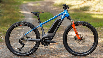 On voit un vélo électrique pour enfants Cube gris, avec des détails bleus et orange. Il se trouve sur un chemin forestier.