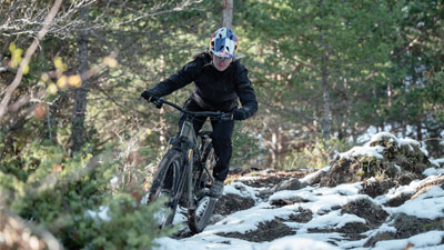 Une cycliste roule à VTT dans une forêt accidentée, où le sol est encore partiellement recouvert de neige.