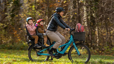 On voit une mère sur un vélo cargo bleu Cube Longtail Hybrid. Deux enfants en bas âge sont assis sur le porte-bagages.